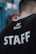 A person wearing a black t-shirt with 'STAFF' written in large white letters on the back. Above it, there is a smaller logo with the words 'Northern Spirit'. The person's head is turned away from the camera, showing only the back of their head and upper body. The background is blurred, with hints of blue and red colors.