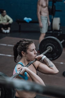A woman is lifting a barbell in a gym setting, focusing intently on her form. She appears to be mid-exercise with a determined expression. In the background, a shirtless man and another person sitting in workout attire are visible. Gym equipment like weights and benches are scattered around.