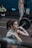 A woman is lifting a barbell in a gym setting, focusing intently on her form. She appears to be mid-exercise with a determined expression. In the background, a shirtless man and another person sitting in workout attire are visible. Gym equipment like weights and benches are scattered around.