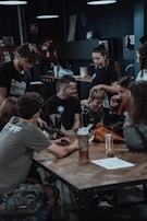 Several people are gathered around a wooden table in a gym-like setting. They appear to be engaged in conversation or looking at something together. There are various items on the table, including a water bottle and a can of an energy drink. The room is dimly lit with overhead lights and some gym equipment and posters are visible in the background.