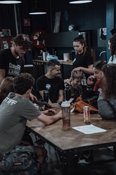 Several people are gathered around a wooden table in a gym-like setting. They appear to be engaged in conversation or looking at something together. There are various items on the table, including a water bottle and a can of an energy drink. The room is dimly lit with overhead lights and some gym equipment and posters are visible in the background.