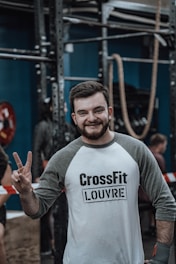 A person is smiling and holding up a peace sign in a gym environment. They are wearing a shirt with the words 'CrossFit Louvre' and are surrounded by gym equipment like ropes and metal bars. Other people are in the background, possibly engaged in activities.