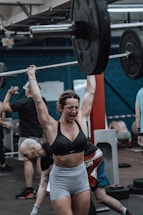 A woman in workout attire is lifting a heavy barbell overhead in a gym environment. Her expression shows intense focus and exertion. The gym appears busy, with other individuals engaged in various exercises in the background.