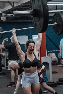 A woman in workout attire is lifting a heavy barbell overhead in a gym environment. Her expression shows intense focus and exertion. The gym appears busy, with other individuals engaged in various exercises in the background.