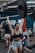 A woman in workout attire is lifting a heavy barbell overhead in a gym environment. Her expression shows intense focus and exertion. The gym appears busy, with other individuals engaged in various exercises in the background.