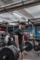 A man is engaged in a deadlift exercise in a gym setting. He is wearing a black t-shirt and appears focused and determined. Several other people are present in the background, all engaged in different fitness activities, creating a lively and energetic environment. The gym is equipped with various fitness gear, including barbells and weights.