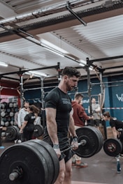 A man is engaged in a deadlift exercise in a gym setting. He is wearing a black t-shirt and appears focused and determined. Several other people are present in the background, all engaged in different fitness activities, creating a lively and energetic environment. The gym is equipped with various fitness gear, including barbells and weights.