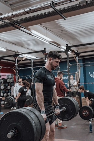 A man is engaged in a deadlift exercise in a gym setting. He is wearing a black t-shirt and appears focused and determined. Several other people are present in the background, all engaged in different fitness activities, creating a lively and energetic environment. The gym is equipped with various fitness gear, including barbells and weights.