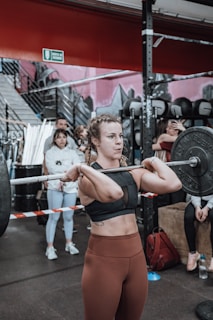 A woman is lifting a barbell in a gym environment. She is wearing a black sports top and brown leggings. There are people in the background observing her lift. The gym has a rugged interior with various equipment visible.