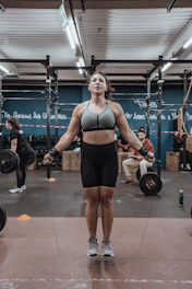 A person is jumping rope in a gym environment while wearing athletic gear. The gym is equipped with exercise equipment such as barbells and ropes. There are other people in the background engaging in fitness activities.