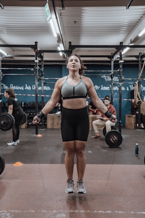 A person is jumping rope in a gym environment while wearing athletic gear. The gym is equipped with exercise equipment such as barbells and ropes. There are other people in the background engaging in fitness activities.