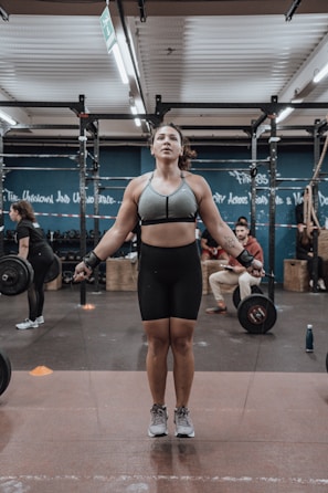 A person is jumping rope in a gym environment while wearing athletic gear. The gym is equipped with exercise equipment such as barbells and ropes. There are other people in the background engaging in fitness activities.