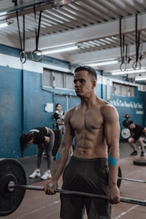 A muscular individual lifting a barbell in a gym setting, showcasing focus and strength. Several people are in the background, engaged in various exercises. The gym features blue and white walls with fluorescent lighting and gymnastic rings hanging from the ceiling.