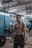 A muscular individual lifting a barbell in a gym setting, showcasing focus and strength. Several people are in the background, engaged in various exercises. The gym features blue and white walls with fluorescent lighting and gymnastic rings hanging from the ceiling.