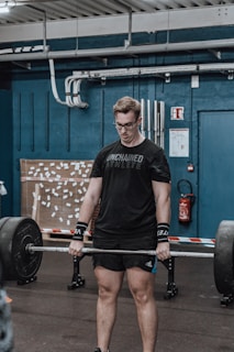 A person is lifting a barbell in a gym setting, displaying focus and determination. The gym environment includes industrial-style elements like exposed piping and a fire extinguisher.