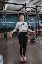 A woman is engaged in a workout, skipping rope inside a gym. She is wearing a white cropped shirt and black leggings, and appears focused on her exercise. In the background, people are involved in various fitness activities, with equipment like kettlebells and a barbell visible. Inspirational words are written on the blue walls of the gym.