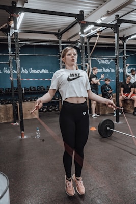 A woman is engaged in a workout, skipping rope inside a gym. She is wearing a white cropped shirt and black leggings, and appears focused on her exercise. In the background, people are involved in various fitness activities, with equipment like kettlebells and a barbell visible. Inspirational words are written on the blue walls of the gym.