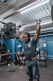 A man is performing a weightlifting exercise in a gym, lifting a heavy barbell above his head. He is wearing a belt and wrist wraps, indicating the intensity of the workout. Several people are sitting in the background watching, suggesting a competition or event setting. The gym is equipped with various fitness equipment, and there is a barrier tape behind the man.
