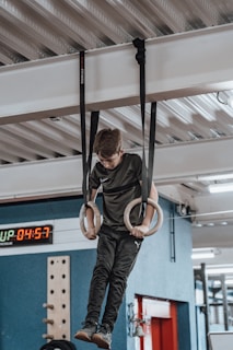 A young person is hanging from a set of gymnastic rings suspended from a ceiling. The setting appears to be an indoor gym with equipment visible in the background, including a digital timer showing the time 04:57.