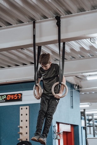 A young person is hanging from a set of gymnastic rings suspended from a ceiling. The setting appears to be an indoor gym with equipment visible in the background, including a digital timer showing the time 04:57.