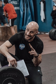 A person with a bald head and a beard is crouching and smiling, pointing a finger upwards. They are wearing a black t-shirt with 'French Throwdown 2024' written on it. A weight plate is visible in the foreground, and other people are exercising in the background in a gym setting.