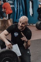 A person with a bald head and a beard is crouching and smiling, pointing a finger upwards. They are wearing a black t-shirt with 'French Throwdown 2024' written on it. A weight plate is visible in the foreground, and other people are exercising in the background in a gym setting.