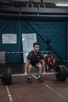 A person is performing a deadlift in a gym setting, surrounded by weightlifting equipment. The walls are painted a dark blue, and gymnastic rings hang from the ceiling. There are leaderboards and instructional boards on the walls.