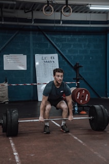 A person is performing a deadlift in a gym setting, surrounded by weightlifting equipment. The walls are painted a dark blue, and gymnastic rings hang from the ceiling. There are leaderboards and instructional boards on the walls.