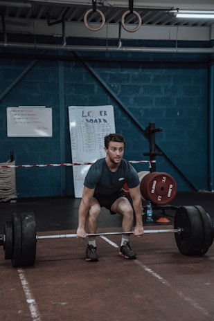 A person is performing a deadlift in a gym setting, surrounded by weightlifting equipment. The walls are painted a dark blue, and gymnastic rings hang from the ceiling. There are leaderboards and instructional boards on the walls.