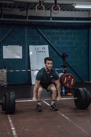 A person is performing a deadlift in a gym setting, surrounded by weightlifting equipment. The walls are painted a dark blue, and gymnastic rings hang from the ceiling. There are leaderboards and instructional boards on the walls.