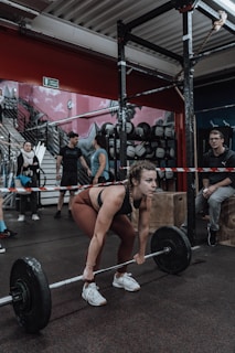 A woman is preparing to lift a barbell in a gym setting. She wears athletic clothing and appears focused. The gym has a variety of equipment including weights and benches, and there are other people interacting in the background, creating a dynamic and energetic atmosphere.