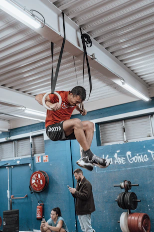 A man in a red shirt is exercising on gymnastic rings suspended from the ceiling in a gym setting. He appears focused and is performing a muscle-up. Two other people are present in the background; one is sitting on the floor looking at a phone, and another is standing while also using a smartphone. The gym has a blue color theme with visible workout equipment such as weights and a fire extinguisher.