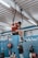A man in a red shirt is exercising on gymnastic rings suspended from the ceiling in a gym setting. He appears focused and is performing a muscle-up. Two other people are present in the background; one is sitting on the floor looking at a phone, and another is standing while also using a smartphone. The gym has a blue color theme with visible workout equipment such as weights and a fire extinguisher.