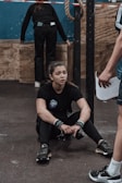 A young woman is seated on the gym floor, appearing exhausted. She holds her knees close, wearing black athletic wear with wristbands. A person in shorts stands nearby, possibly a coach or trainer, holding a piece of paper. Another person is in the background, facing away and interacting with gym equipment. A rope is hanging from the ceiling and wooden boxes are visible.
