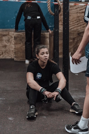 A young woman is seated on the gym floor, appearing exhausted. She holds her knees close, wearing black athletic wear with wristbands. A person in shorts stands nearby, possibly a coach or trainer, holding a piece of paper. Another person is in the background, facing away and interacting with gym equipment. A rope is hanging from the ceiling and wooden boxes are visible.