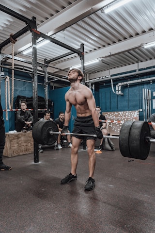 A man is performing a deadlift in a gym setting, surrounded by onlookers who appear to be observing or monitoring his performance. The gym is equipped with various fitness equipment, including a pull-up bar and stacked weights. The atmosphere suggests a communal workout environment.