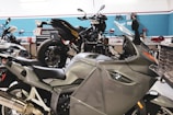 A workshop with multiple motorcycles, including a prominent silver BMW motorcycle in the foreground. The space is organized with tools and equipment along the walls and workbenches.