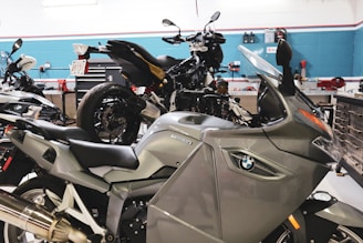 A workshop with multiple motorcycles, including a prominent silver BMW motorcycle in the foreground. The space is organized with tools and equipment along the walls and workbenches.