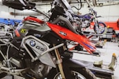 A well-equipped motorcycle workshop featuring a prominently displayed BMW GS motorcycle with red and black colors. The bike is mounted on a lift with various tools and equipment in the background. Another motorcycle can be seen on a lift in the background, highlighting a professional garage setting with organized workspaces.