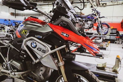 A well-equipped motorcycle workshop featuring a prominently displayed BMW GS motorcycle with red and black colors. The bike is mounted on a lift with various tools and equipment in the background. Another motorcycle can be seen on a lift in the background, highlighting a professional garage setting with organized workspaces.