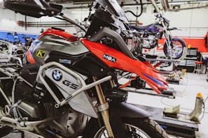 A well-equipped motorcycle workshop featuring a prominently displayed BMW GS motorcycle with red and black colors. The bike is mounted on a lift with various tools and equipment in the background. Another motorcycle can be seen on a lift in the background, highlighting a professional garage setting with organized workspaces.