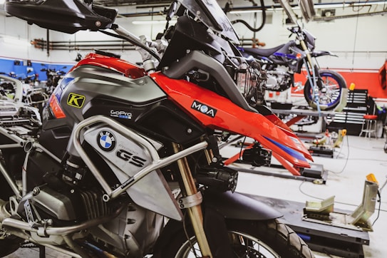 A well-equipped motorcycle workshop featuring a prominently displayed BMW GS motorcycle with red and black colors. The bike is mounted on a lift with various tools and equipment in the background. Another motorcycle can be seen on a lift in the background, highlighting a professional garage setting with organized workspaces.
