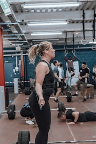 Several people are engaged in various fitness activities inside a gym. A woman in the foreground is exercising with a jump rope, while others are lifting weights and performing exercises in the background. The gym has a high ceiling with exposed pipes and industrial lighting.