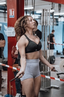 A woman is engaged in a jump roping exercise in a gym setting. She has curly hair and is wearing a sports bra and shorts. The gym is busy with other people, some exercising and others watching. Gym equipment like weights and ropes are visible in the background.