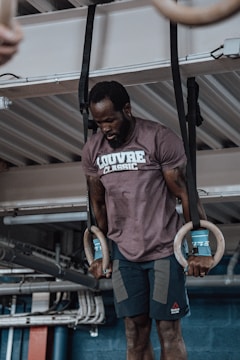 A man is exercising on gymnastic rings in an industrial setting. He is wearing a brown t-shirt with the text 'LOUVRE CLASSIC' and dark shorts. The background shows metal beams and pipes, suggesting an indoor gym environment. The man appears focused and is using wrist supports while holding the rings.