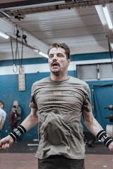 A man is jumping rope in an indoor gym setting. He is wearing a green Reebok CrossFit shirt and black wrist wraps. Behind him, there are gym equipment and another person visible. The gym has a blue and white color scheme with fluorescent lighting.