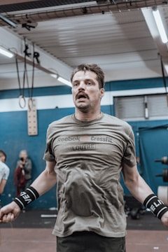 A man is jumping rope in an indoor gym setting. He is wearing a green Reebok CrossFit shirt and black wrist wraps. Behind him, there are gym equipment and another person visible. The gym has a blue and white color scheme with fluorescent lighting.