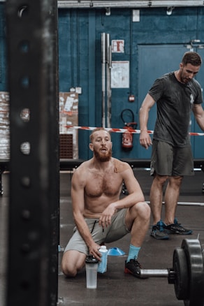 Two men are in a gym setting, one is kneeling with a shaker bottle in hand and looking up, appearing tired. The other man stands behind him, also looking fatigued, wearing athletic clothing. Exercise equipment like weights can be seen around.