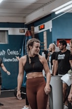 A young woman with braided hair is in a gym setting, wearing a black sports bra and brown leggings. She appears to be conversing with or listening to people around her. The background shows people engaged in a fitness environment, with gym equipment and motivational chalk writings on the wall.