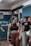 A young woman with braided hair is in a gym setting, wearing a black sports bra and brown leggings. She appears to be conversing with or listening to people around her. The background shows people engaged in a fitness environment, with gym equipment and motivational chalk writings on the wall.